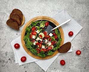 Greek salad with tomatoes, cucumbers, olives, feta cheese, herbs in a plate, fork, bread. Light background. Top view, flat lay.