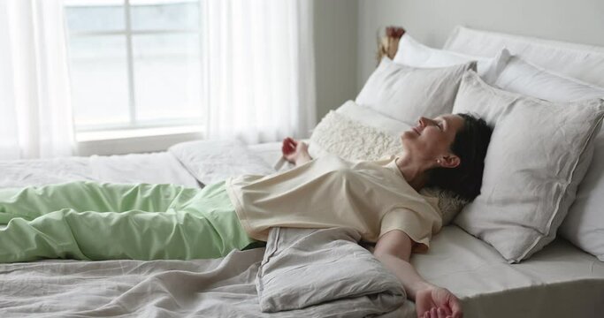Cheerful excited middle aged older woman falling down in bed, throwing body on mattress, linen, resting in bedroom, hotel room, closing eyes, smiling, enjoying stress relief, relaxation