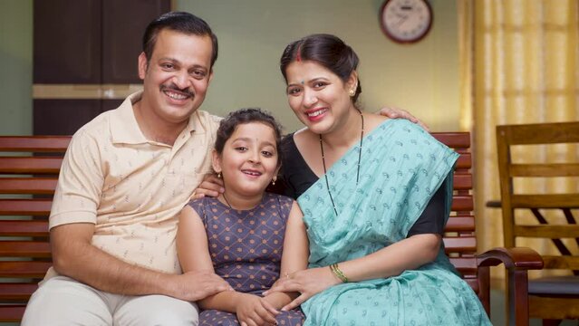 Happy smiling couple with daughter looking camera while sitting on sofa at home - concept of relationship, family photo posing and togetherness