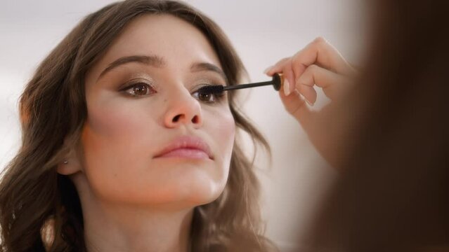 Young woman uses wand to apply black long lasting mascara on eyelashes for volume. Attractive lady with curly hair does makeup for graduation party closeup