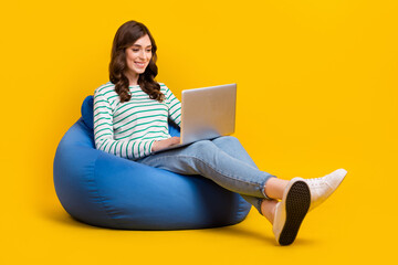 Full size photo of positive cheerful woman striped sit on armchair look at laptop typing email isolated on yellow color background