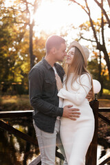 Happy stylish pregnant woman and man hugs, kiss, touches belly and listen baby's kicks, walk outdoor in autumn park. Pregnancy, motherhood and parenthood concept. Family is expecting for unborn baby.