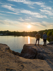 Aerial drone shot of the silhouettes of two people father and son watching the dramatic and colorful sunset with orange, yellow and grey clouds over the trees on the shore of peaceful forest lake