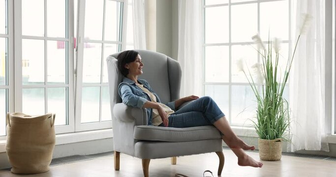 Happy pretty mature lady resting in comfortable home armchair at big windows, listening to relaxing music, enjoying leisure, break, being in cozy warm apartment interior. Side full length shot