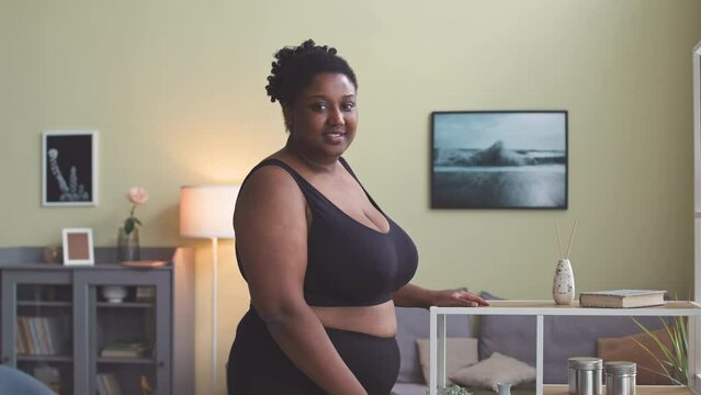 Medium portrait of young plus size African American woman in tight black activewear posing for camera standing in bright modern living room