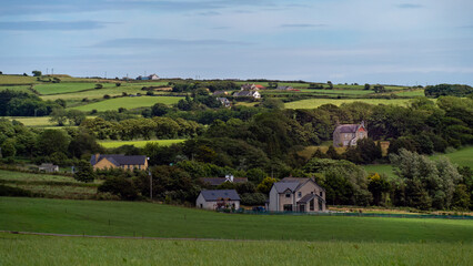 Fototapeta na wymiar A European village among green fields, trees. Farmhouses in Ireland. Picturesque landscape. Green fields under a blue sky, houses on green grass field.