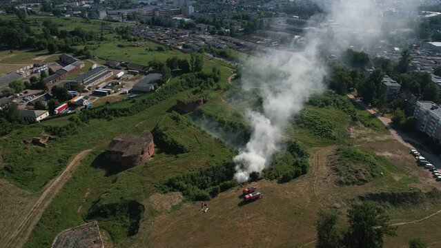 Top view of fire trucks near column of smoke in wasteland next to old fortress across road from residential buildings