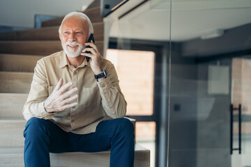 Happy relaxed senior man wearing beige shirt and hand watch talking on mobile phone while sitting...