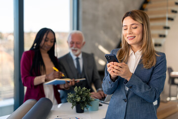 Smart confident business woman company female manager talking using mobile phone technology at work place. Two business persons diverse business team working in background writing ideas in notebook