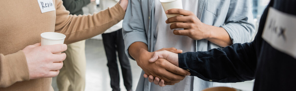 Cropped view of multiethnic people with alcohol addiction holding paper cups and shaking hands in rehab center, banner.