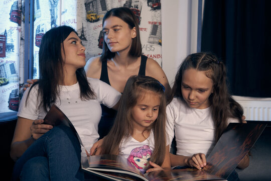 Staged photo. Lesbian couple and their kids are having a good time at home.  Parents and girls are nestled on the couch. The girls are looking at a picture book. Parents gazing tenderly at each other.