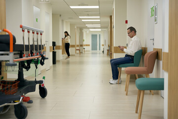 Fototapeta premium Man in glasses sits in a hospital corridor with a phone in his hands