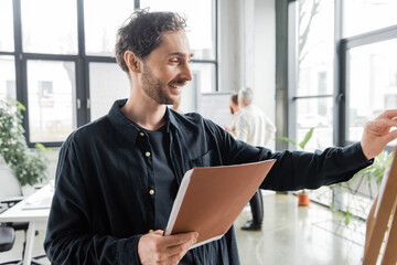 Cheerful businessman with paper folder working with board in office.