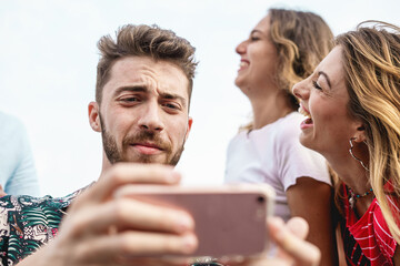 Young man looking troubled at his phone while female friends joke around him