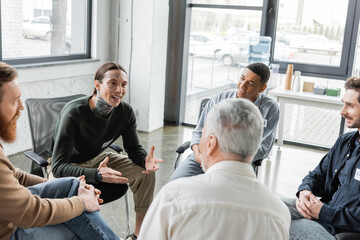 Cheerful tattooed man with alcohol addiction talking to interracial group during therapy in rehab...
