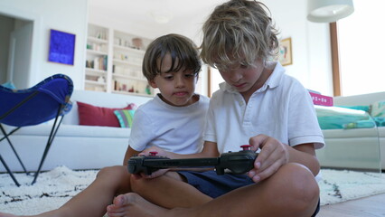 Children looking at video game screen tablet. Younger brother watching sibling play games on joystick console. Kids playing with technological device at home living room © Marco