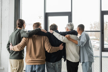 Back view of interracial group of anonymous alcoholics hugging during meeting in rehab center.