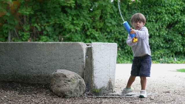 Child getting water from fountain and spraying with foam blaster toy gun outdoors