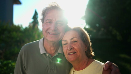A happy married older couple looking at camera at park with sunlight flare backlight. Close up of senior faces of 70s husband and wife