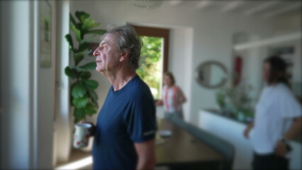 A contemplative senior man standing at home living room while drinking coffee. Mature older person staring out window