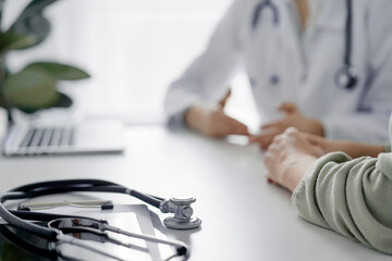 Doctor and patient are sitting and discussing something at the desk in the clinic office. The focus is on the stethoscope lying on the table, close up. Perfect medical service and medicine concept.