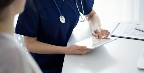 Doctor and patient sitting at the table in clinic while using tablet computer. The focus is on female physician's hands, close up. Medicine concept.