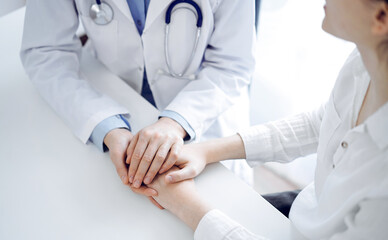 Doctor and patient sitting near each other at the table in clinic office. The focus is on female physician's hands reassuring woman, only hands, close up. Medicine concept.