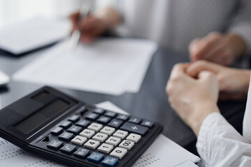 Woman accountant using a pen and laptop computer while counting and discussing taxes with a client, focus on the calculator. Business audit and finance concepts.