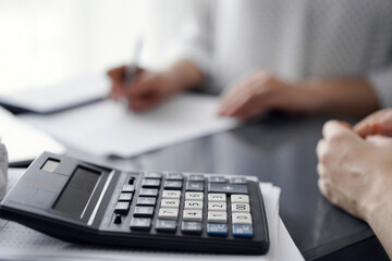 Woman accountant using a pen and laptop computer while counting and discussing taxes with a client, focus on the calculator. Business audit and finance concepts.