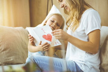 Happy mother day in sunny flat. Child daughter congratulates mom and gives her postcard with heart drawing. Family concept