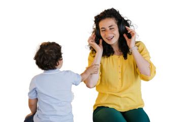 Laughing curly hispanic young adult woman in yellow blouse and green pants sitting against transparent background with little son listening music eyes closed using headphones. Mom and son having fun