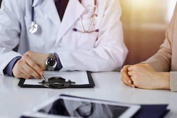 Fototapeta premium Unknown male doctor and patient woman discussing something while sittingin a darkened clinic and using clipboard, glare of light on the background