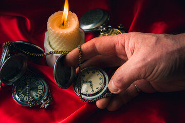 man's hand holds a vintage pocket watch, in the background a burning candle on a red background....