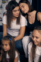 Staged photo. A lesbian couple and their children are having a good time at home.  While the kids are watching a cartoon, the parents are cuddling affectionately.