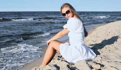 Fototapeta na wymiar Happy, beautiful blonde woman is sitting on the ocean beach in a white summer dress and sunglasses.