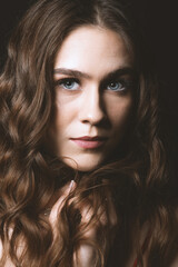 Fototapeta premium Beautiful brunette woman with long wavy hair and blue eyes close-up studio portrait. Model holding hair in hands and looking to camera with serious look. Toned image with soft beige color