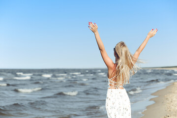 Happy blonde beautiful woman on the ocean beach standing in a white summer dress and glasses, raising hands.