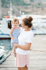 Fototapeta na wymiar Mother holding baby girl 1-2 year old wearing casual clothes walking at boat pier over sea background outdoor. Motherhood. Summer vacation season.