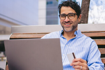 Middle-aged man looking at camera while holding a credit card in his hand and his laptop on his lap.