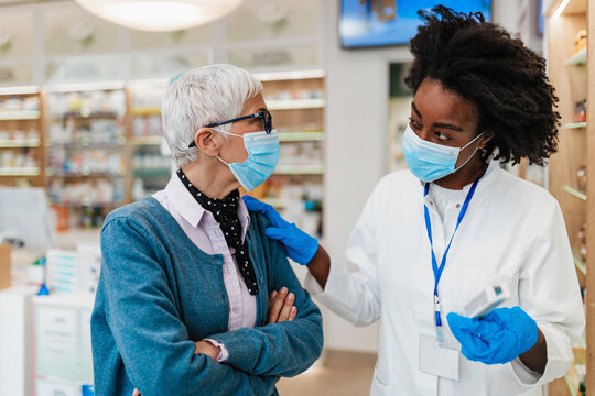 Professional Black Female Pharmacist Talking With Senior Customer In Modern Drugstore. They Are Wearing Face Protective Masks For Protection From Coronavirus.