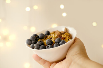 close-up female hand holds healthy food white cup, oatmeal, blueberries, nuts, almonds, peanuts, raisins, dried fruits on blurred background with bokeh, raw food diet, vegetarian food