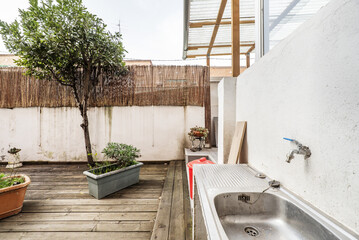 Terraced patio of a ground floor apartment with some plants and unvarnished acacia wood floors, a stainless steel sink, a partition and a small tree