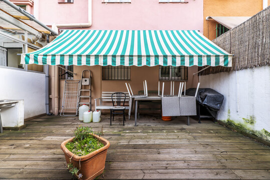 Terraced Courtyard Of A Ground Floor Apartment With Some Plants And Unvarnished Acacia Wood Floors, Two-tone White And Green Extended Fabric Awning And Stacked Furniture