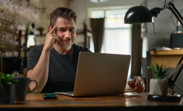 Bearded man working online with laptop computer at home sitting at desk. Home office, browsing internet. Portrait of mature age, middle age, entrepreneur.