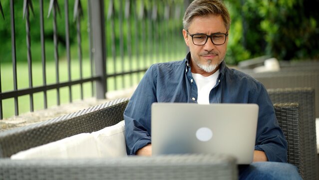 Man sitting on outdoor sofa in garden working with laptop computer in home office, managing business online, thinking