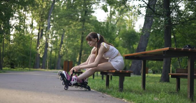 A girl on roller skates sits on a bench in a park full of trees