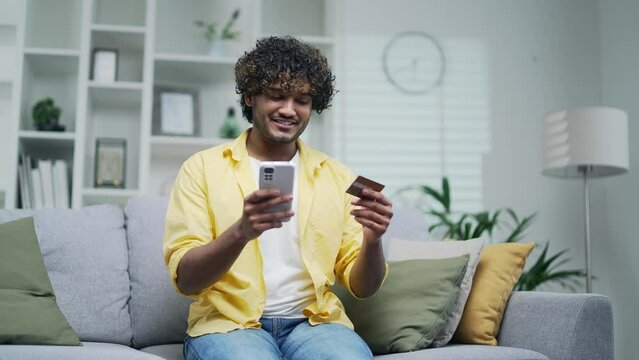 Young adult man pay for services makes a payment using smartphone and credit card, sitting at home on sofa. A happy male is using a mobile app booking orders by phone Makes purchases in online stores