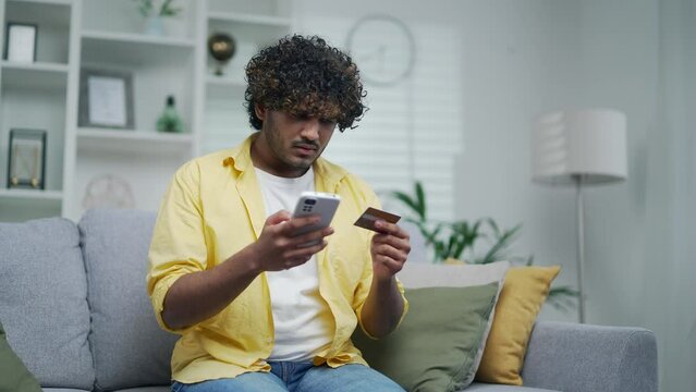 Unsuccessful payment internet scam Fraud victim Upset indian guy sit on sofa at home holds card and smartphone phone palm feels shocked, overspending money lost savings money stolen from bank problem