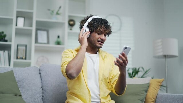 Cheerful young man in a bright yellow shirt listens to music in headphones using app application in a mobile smartphone phone Handsome male Enjoys and relaxes sitting at home on the sofa.Indoor dances