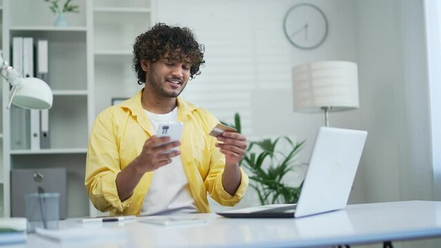 Young adult man pay for services makes a payment using smartphone and credit card, sitting at office or home Happy male using a mobile app booking orders by phone Makes purchases in online stores
